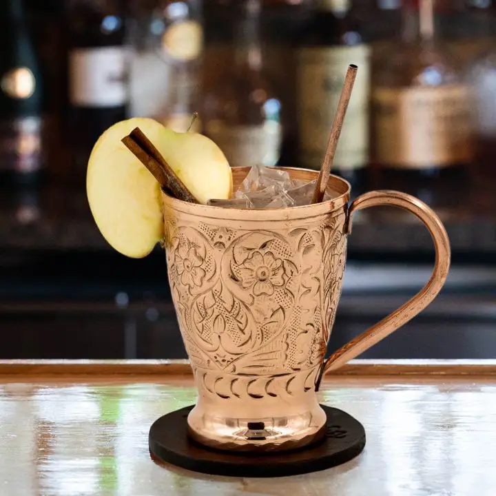 A tapered copper Moscow Mule mug has intricate flower designs on it and a swooping handle. It rests on a leather coaster on a reflective bar top, with ice poking up, a slice of apple and cinnamon stick as garnish. 