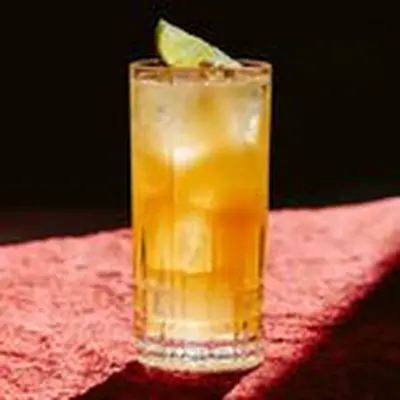 A faceted Collins glass holds a red-orange drink garnished with a lime wedge. It sits on a red stone surface with dramatic lighting.