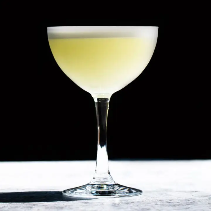 A brightly lit cocktail coupe holds a vidid yellow cocktail with a white foamy head. The glass stands on a white stone surface, with a jet black background. 