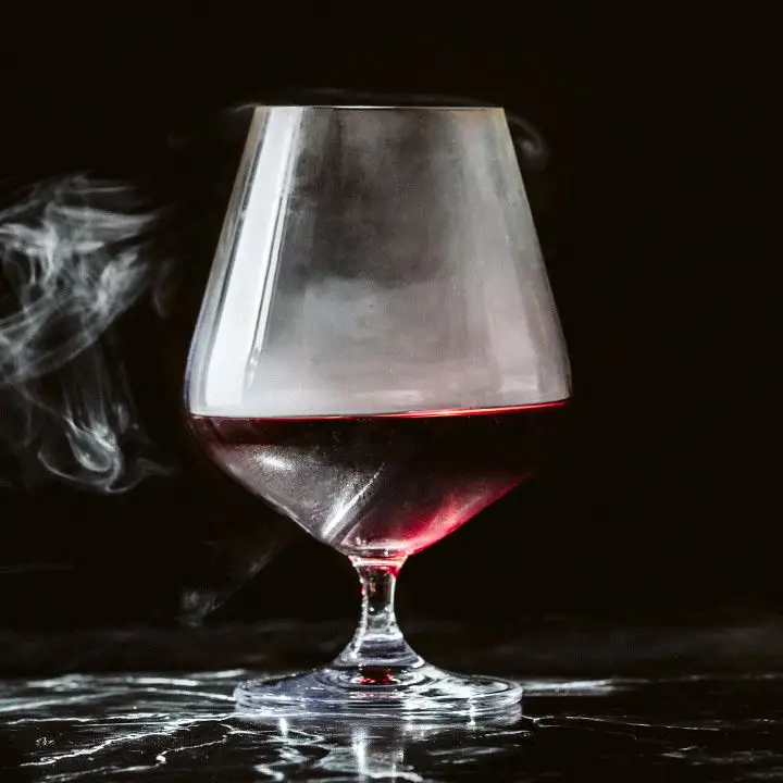 A large snifter rests on a black marble surface against a sold black backdrop. The snifter contains a dark red drink and whiffs of smoke which emanate from the glass. 