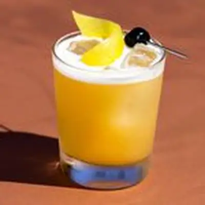 Amaretto Sour in rocks glass with egg white foam on top, garnished with lemon twist and maraschino cherry