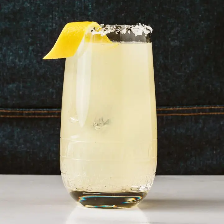 A Collins glass rests on a white surface, with a denim apron behind it. The drink is a pale yellow with ice and a salted rim. 
