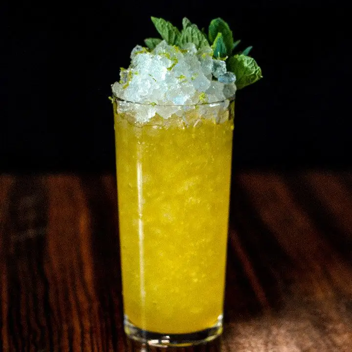 A tall Collins glass is filled with crushed ice and a golden drink, and is garnished with mint leaves. It rests on a dark wooden surface.