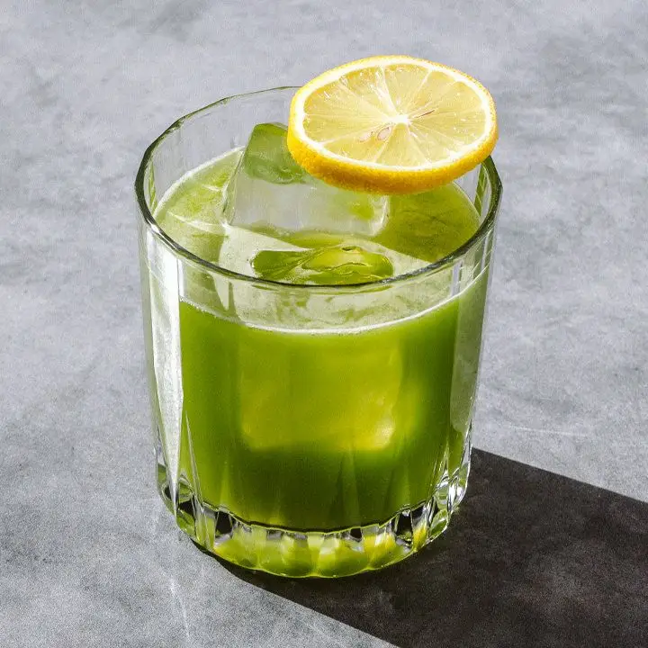 green-colored Green Tox cocktail in a rocks glass with ice cubes and a lemon wheel balanced on the rim, served on a sunny gray surface
