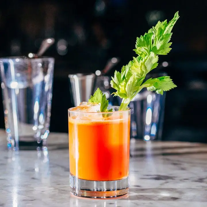 bright-orange Lunar Eclipse cocktail in a rocks glass, garnished with a leafy celery stick and served on a bar in front of glasses and tools