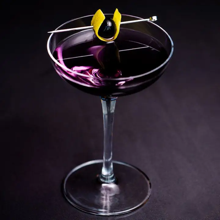 An elegant coupe glass holds a deep violet drink, garnished with a cherry wrapped in a lemon peel, pierce by a pick. The glass sits on a dark gray surface.