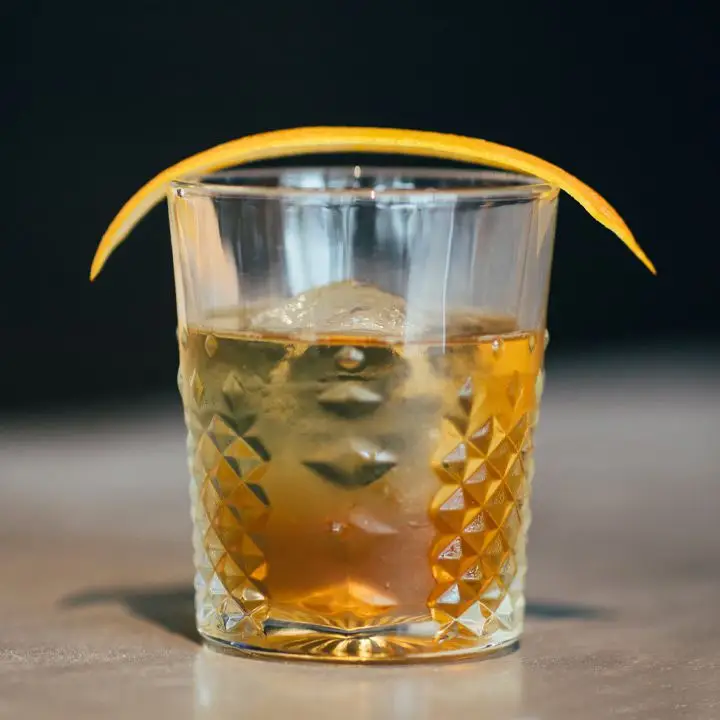 Forefathers cocktail in a textured rocks glass with one large ice cube and a large orange peel balanced on the rim