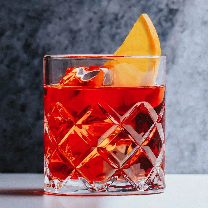 A sturdy looking rocks glass is filled with clear ice and the iconic crimson-hued Negroni, and is garnished with a slice of orange. The background is gray.