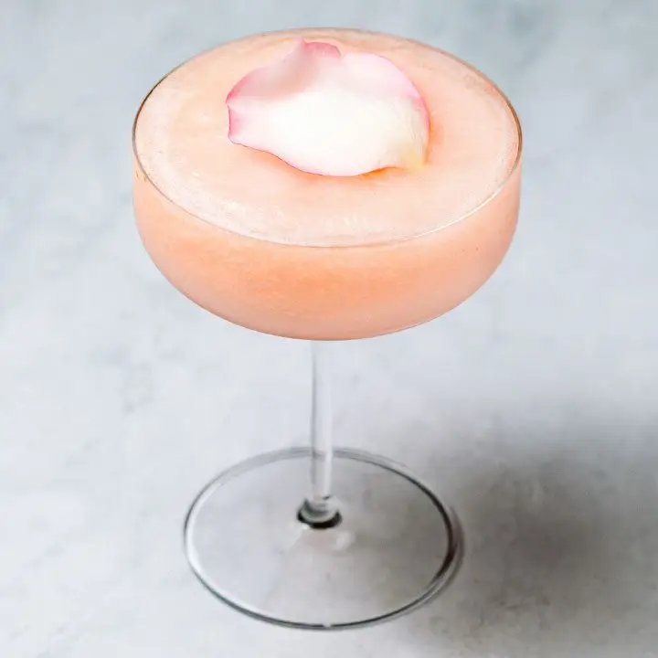 Icy, peach-colored Guava Rose cocktail in a coupe glass, garnished with a pink rose petal