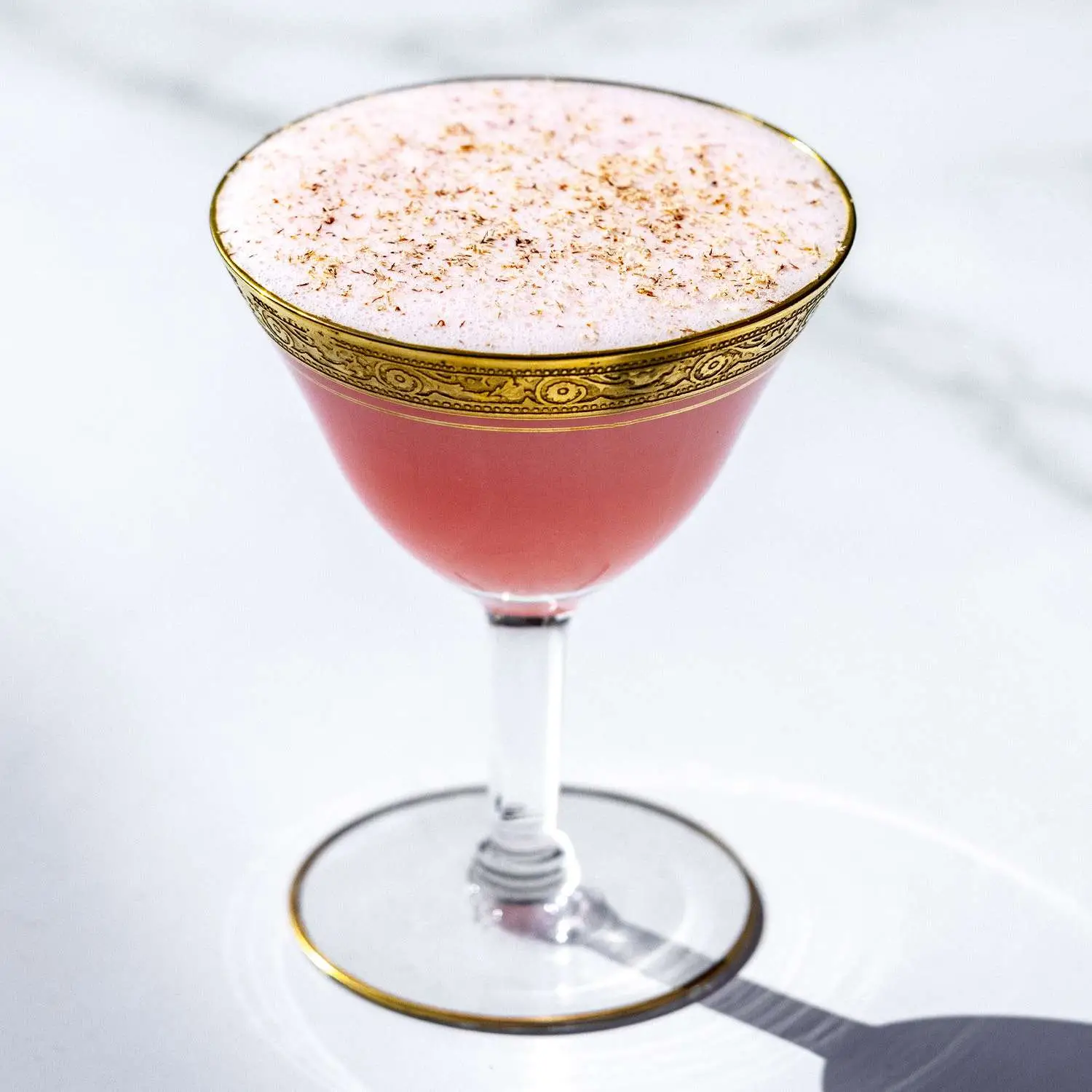 Pink-hued Millionaire cocktail in ornate coupe glass with etched gold rim, dusted with nutmeg, on white marble background 