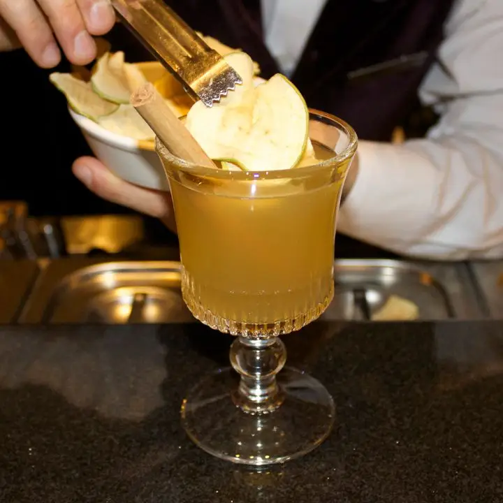 hand places dried apple garnish on the Hot Apple Pie cocktail, served in an Irish Coffee glass with a cinnamon stick