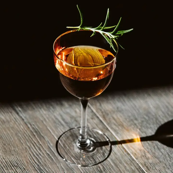 A Nick & Nora glass stands on a dramatically lit wooden surface, the background in shadow. The drink within is a golden hue, garnished with a rosemary sprig.