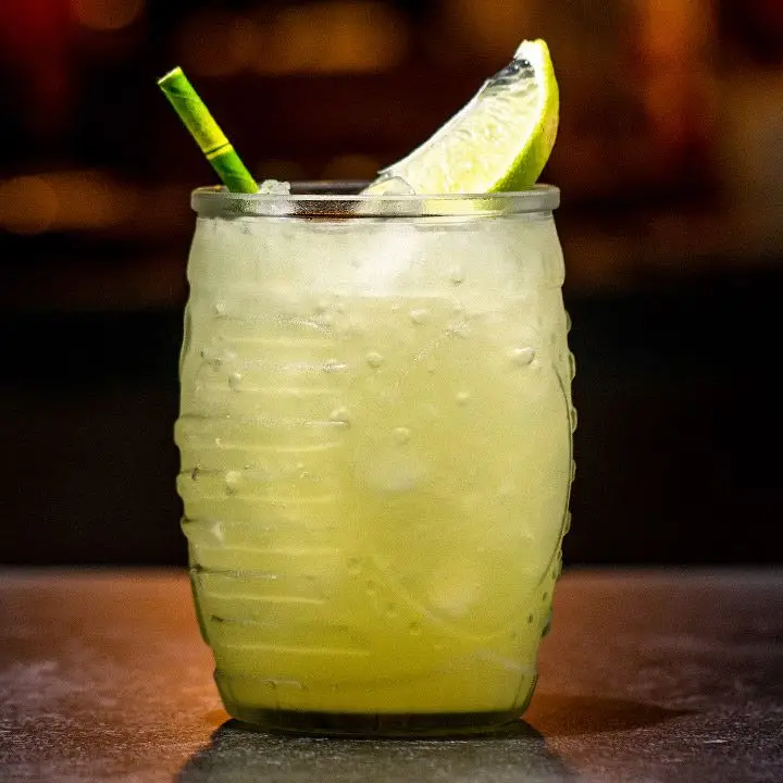 A bright yellow-green cocktail served in a thick barrel-shaped glass with a lime wedge and a green paper straw