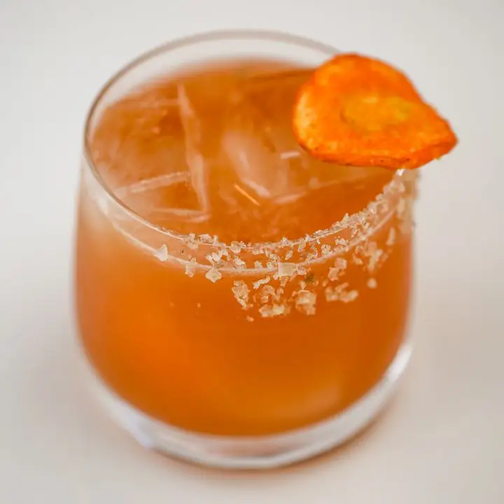 A rocks glass is photographer in tight, shallow focus. The drink within is carrot-orange over a few ice cubes, garnished with a dry carrot ring and flaky salt.