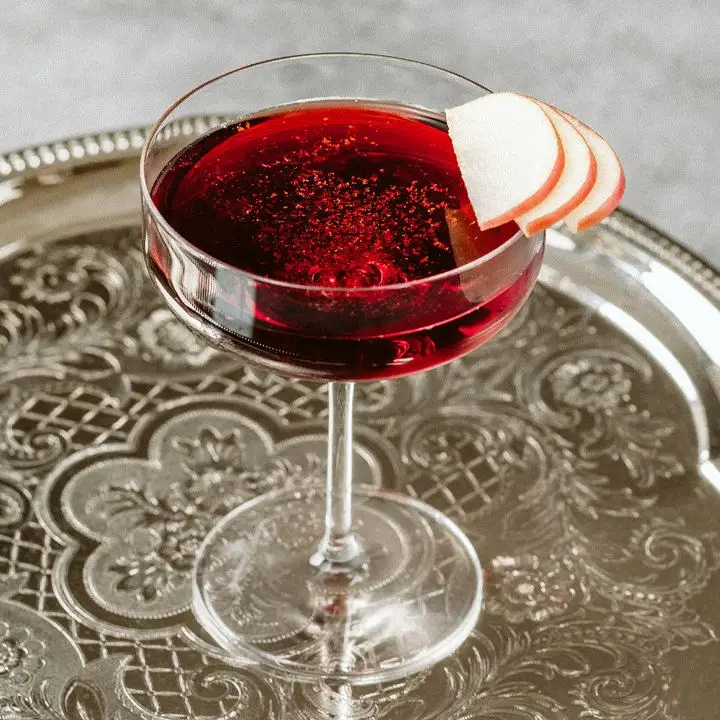 A wide coupe glass rests on a lacquered silver bar tray. The glass is filled with a crimson cocktail and garnished with grated cinnamon and apple slices.