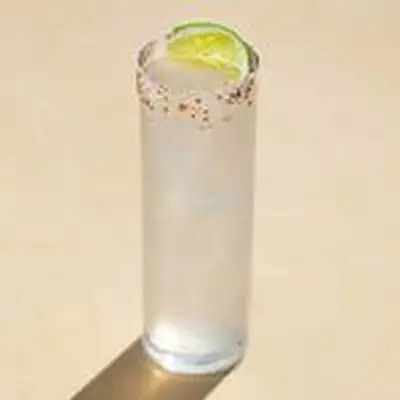 Mezcal Paloma in highball glass with Tajin salt rim and lime wedge, shot against tan background