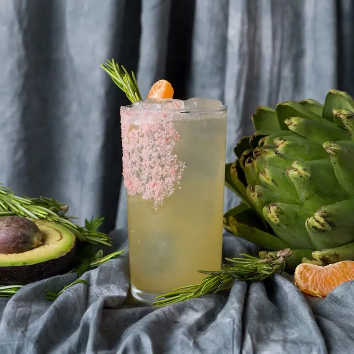 A yellow-hued cocktail in a clear Collins glass with a generous pink salt garnish on the side of the glass and a variety of fruits and vegetables surrounding the drink