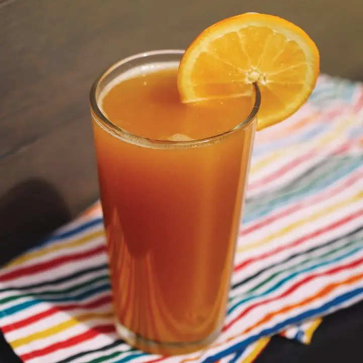 A tall, thin beer glass rests on a colorful striped cloth on a wooden surface. The glass holds an opaque orange drink and is garnished with an orange wheel.