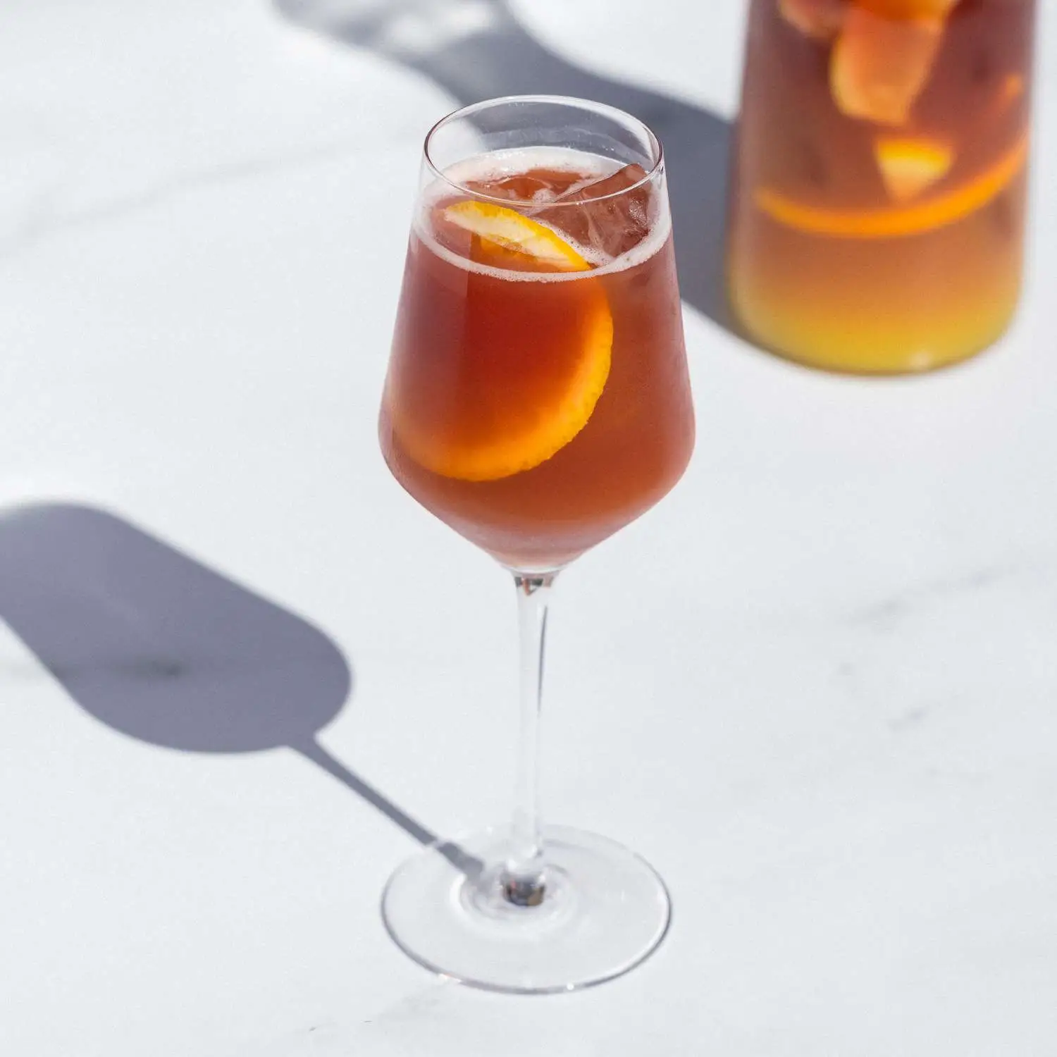 Red Sangria in wine glass with ice and orange slice on white marble, with pitcher of Sangria in background