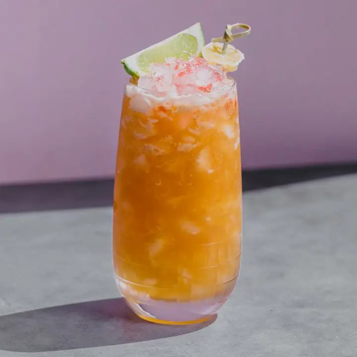 A curved Collins glass rests on a concrete surface with a purple backdrop. The drink within is deep orange, filled with crushed ice and garnished with a lime wedge and bitters.