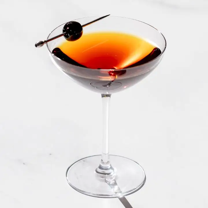 Gotham cocktail in a coupe glass with a skewered cherry balanced on the rim