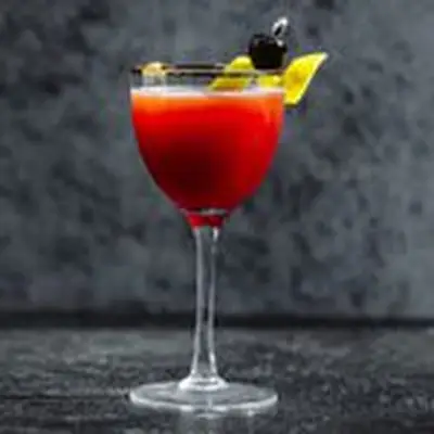 A Nick & Nora with a copper rim rests on a marble gray backdrop. It holds a crimson drink, garnished with lemon peel and cherry.