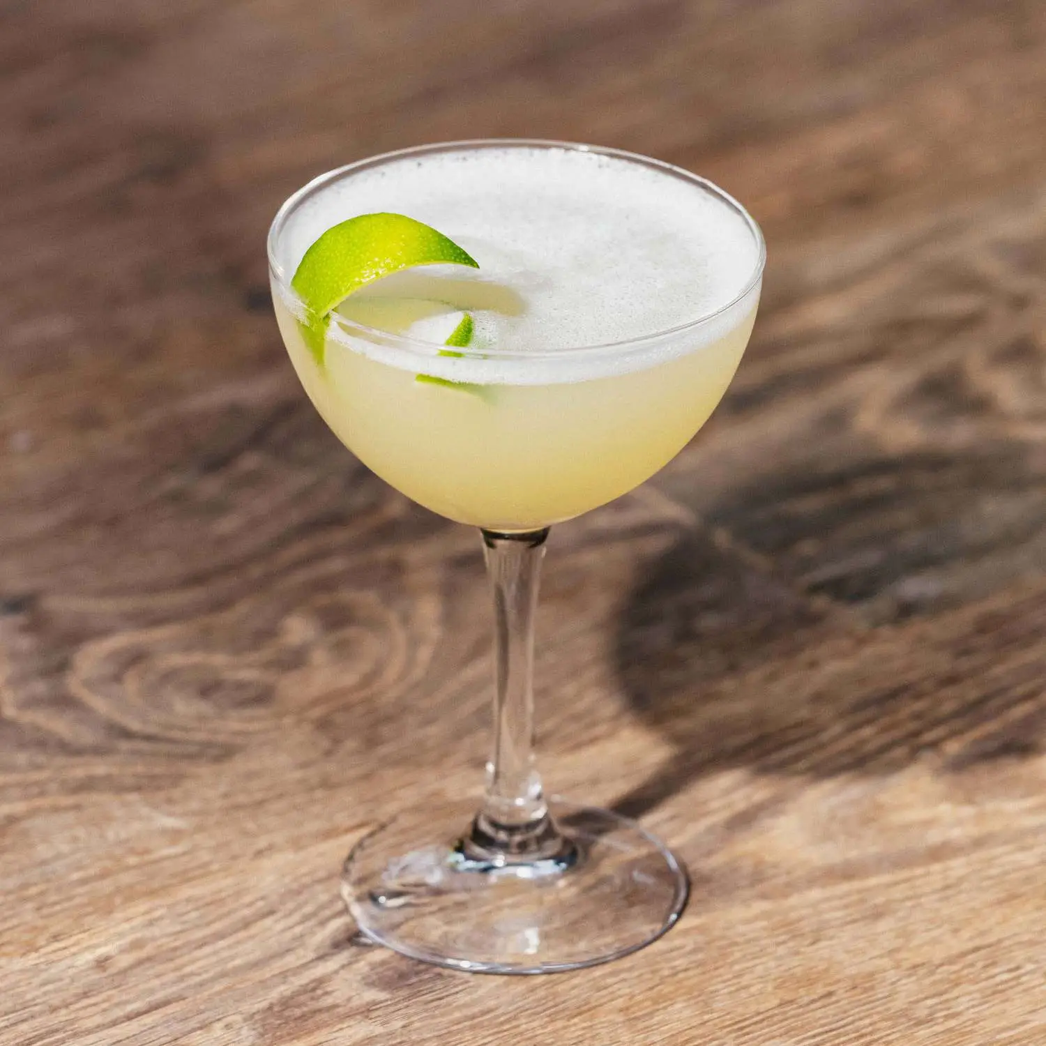Daiquiri in coupe glass with light foam and lime twist on top, on wood counter