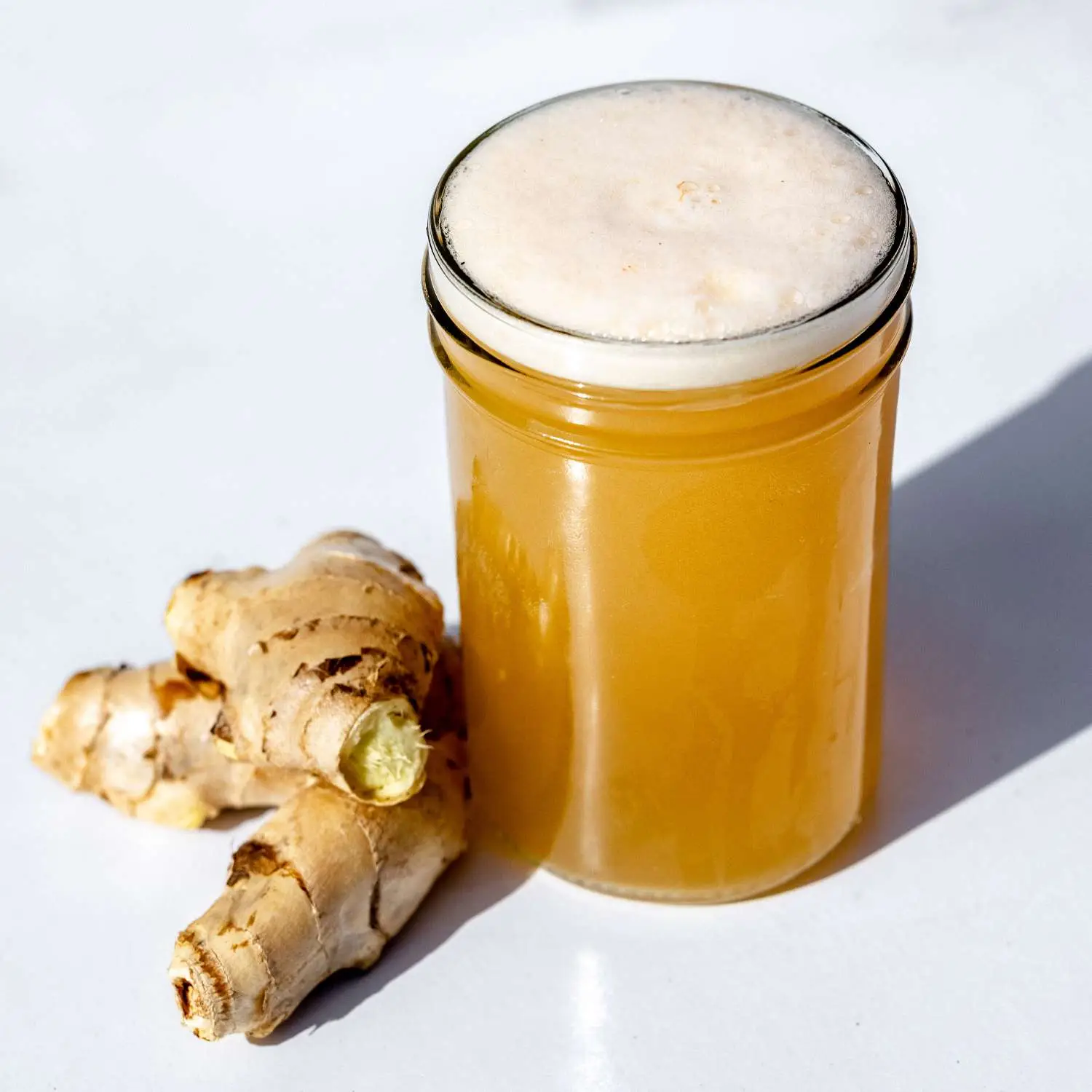 Ginger beer with foamy head, in small tumbler glass seated next to knob of fresh ginger