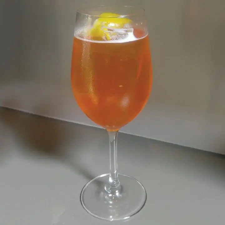 A wine glass stands on a neutral gray background, and is filled with a red-orange bubbly drink. A few ice cubes float in the glass, and it’s garnished with a lemon peel.
