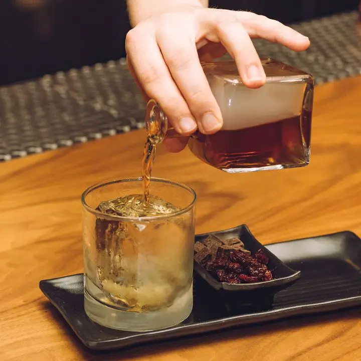 hand pours a small decanter of the Smoke Break cocktail into a rocks glass over one large ice cube