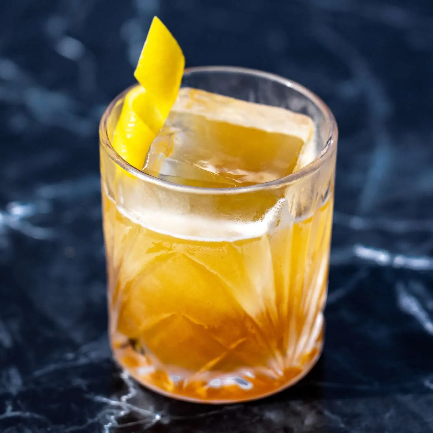 Gold Rush cocktail in an engraved rocks glass with single large ice cup and lemon twist, on a dark marble background