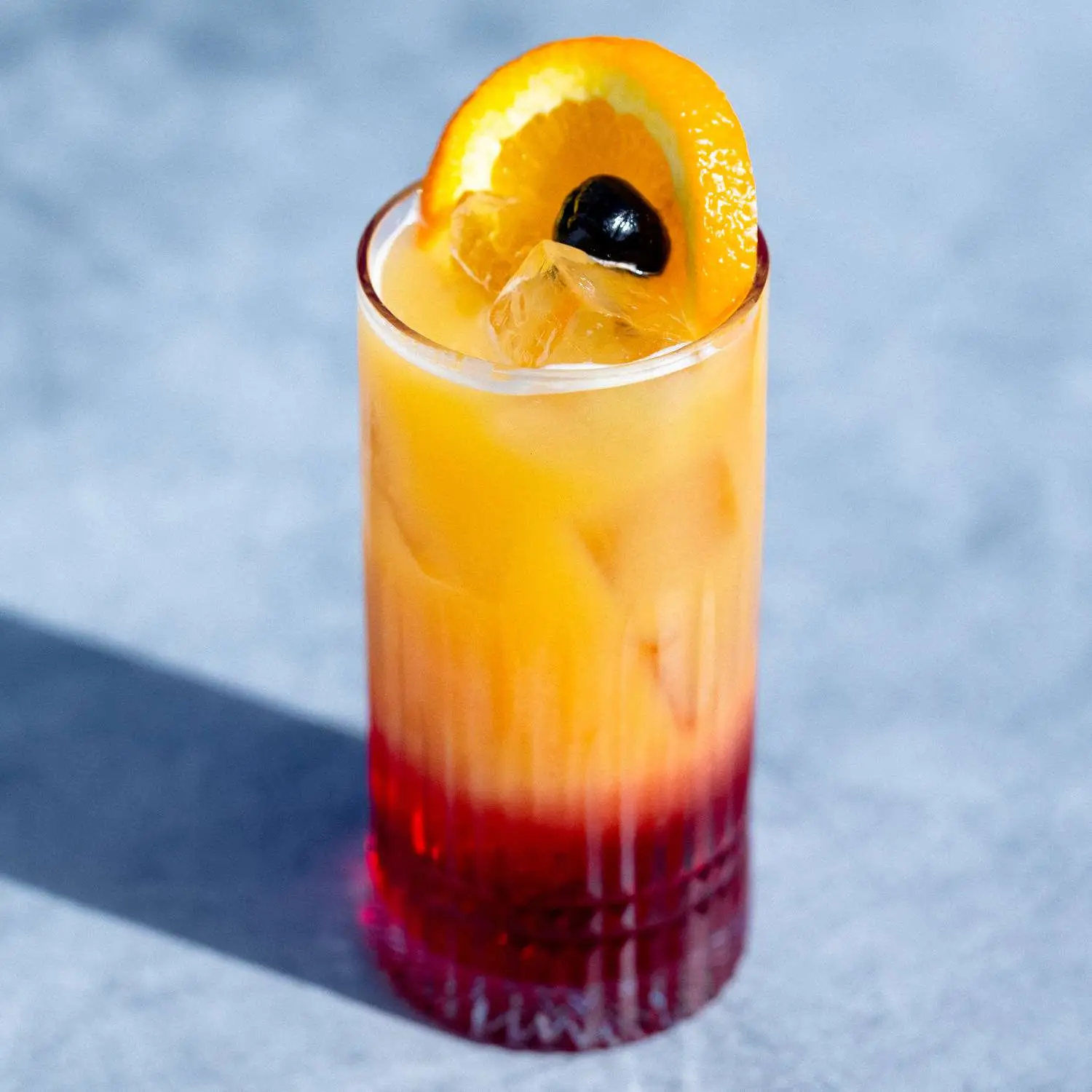 Layered Tequila Sunrise cocktail in highball glass, garnished with maraschino cherry and orange wheel, shot against gray background