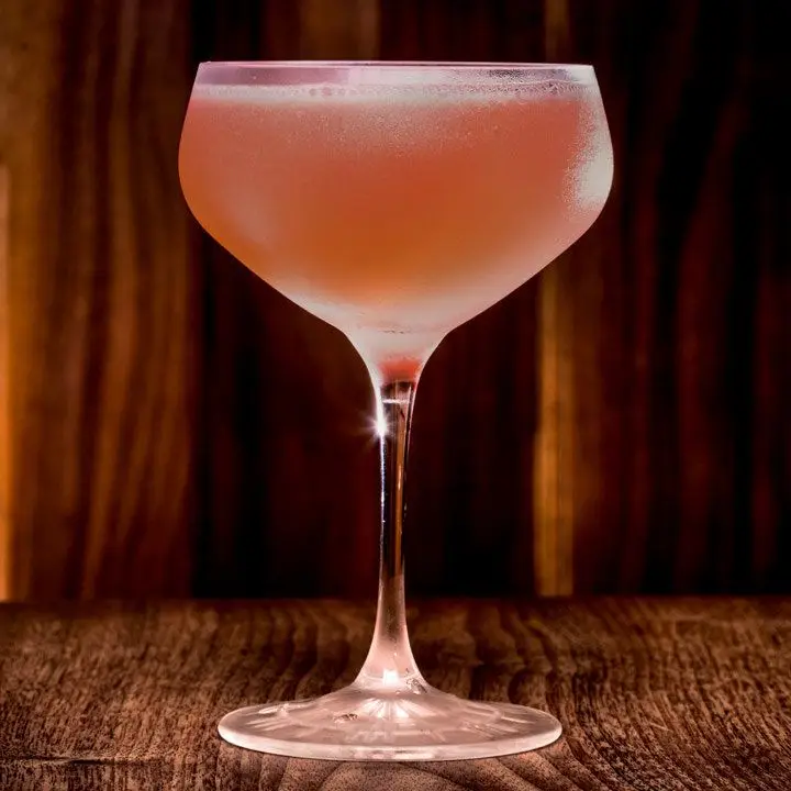 A deep coupe glass rests on a hard wood surface with a wood backdrop. The drink within is dark red-brown.