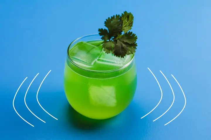 A jade colored cocktail with a few large ice cubes fills a curved rocks glass, topped with a sprig of cilantro. Its background is pure blue with a few emphasis lines