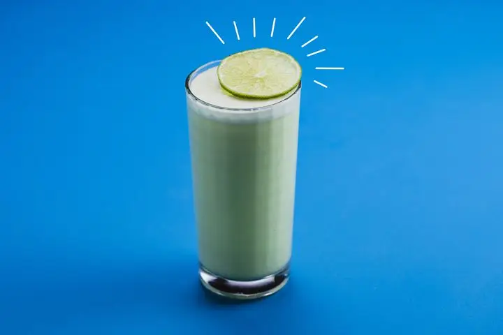 A tall, square-faceted collins glass is filled with an opaque, pastel green beverage, all on a solid blue backdrop. A wheel of lime rests atop the glass, emphasized with straight white lines