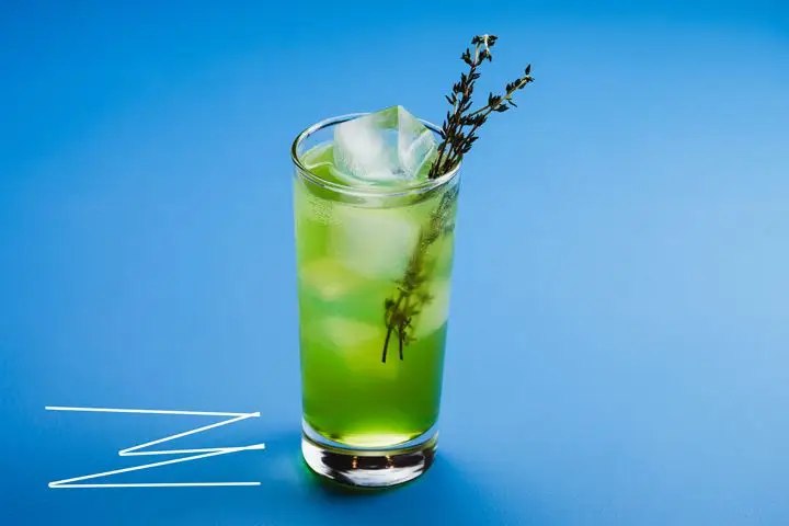 A tall, thin glass holds a few large ice cubes and an emerald green beverage, garnished with two sprigs of thyme. It sets on an all blue backdrop; adjacent are a few zig zagging white lines