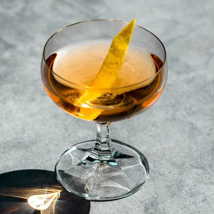 An elegant coupe with a faceted stem rests on smooth gray concrete. The glass holds a light amber drink and a thin slice of lemon.