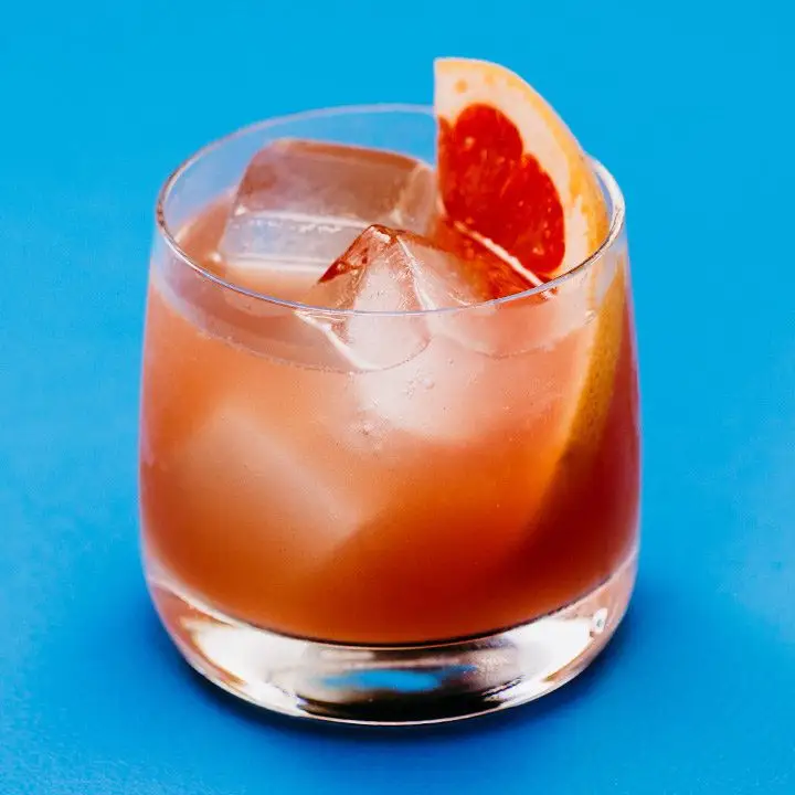 A double Old Fashioned glass is against a solid sky-blue backdrop. The glass is filled with a few large ice cubes, a juicy orange drink and a thin slice of grapefruit.