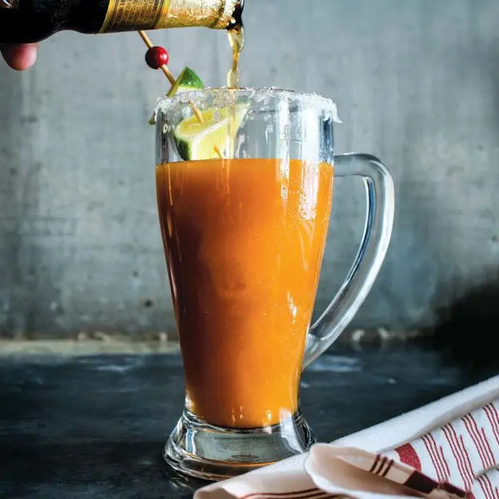 hand pours a dark beer into the orange-colored Shannon’s Sangrita/Michelada, served in a beer mug with skewered lime garnish beside a cloth napkin