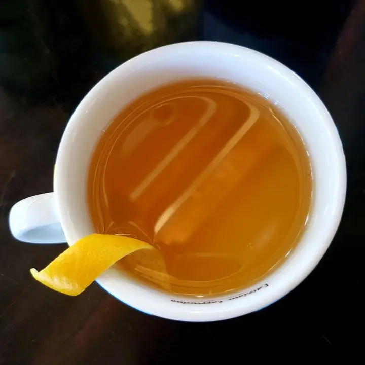 A top down view of a white mug shows a light brown drink garnished with a thin peel of orange. The background is reflective black.