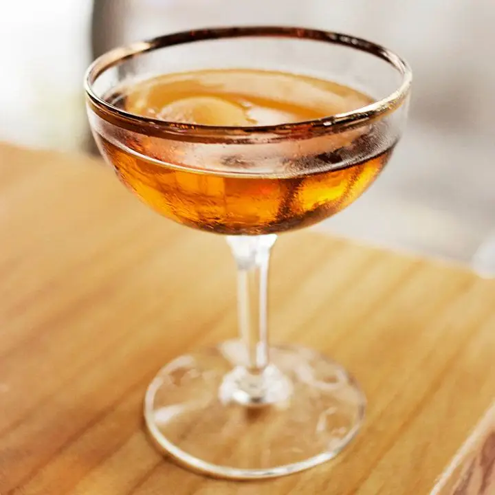 A coupe with a copper rim rests on a light wooden table. It’s filled with a pale gold cocktail.