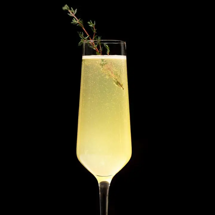 A narrow Champagne flute holds a bubbly yellow drink garnished with a sprig of thyme. It glows brightly against a black background
