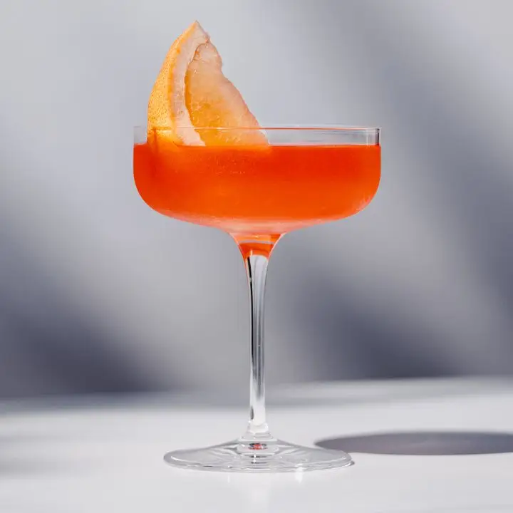 A flat-bottomed coupe on a white surface holds a vivid orange drink garnished with a large slice of grapefruit.