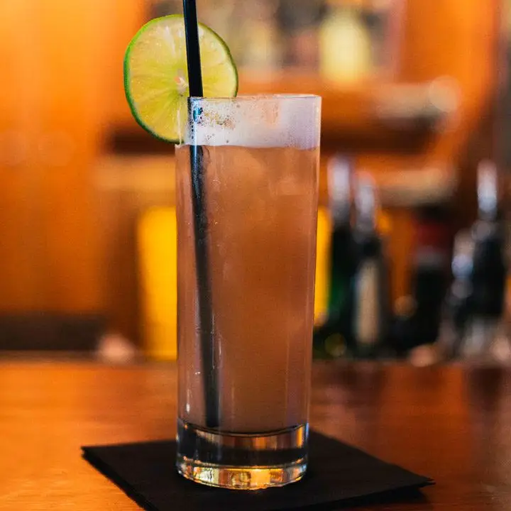 A traditional highball glass rests on a black cocktail napkin on a bar, the background hazy and indistinct. The drink is a dark brown, and garnished with a long black straw and a lime wheel.