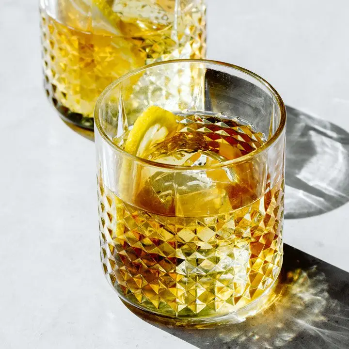 With dramatic lighting casting dark shadows on a brilliantly lit white background, two detailed rocks glasses hold ice, a slice of lemon, and a vivid orange punch.