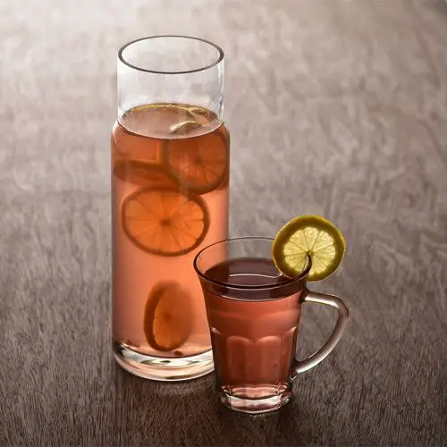 A tall, narrow glass pitcher with no handles is filled with a pale orange-pink drink, in which floats numerous slices of lemon. A glass mug to its right has the same drink and a lemon wheel garnish.