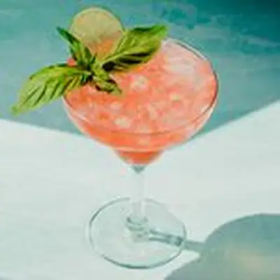 In a busy photo, two short, round glasses are rimmed with rose salt and filled with a bubbly pink punch, strawberries and lime. In the background is the punchbowl with the same drink, and limes and rose salt rest on a cutting board. The background is blue, including a patterned blue and white cloth under one glass.