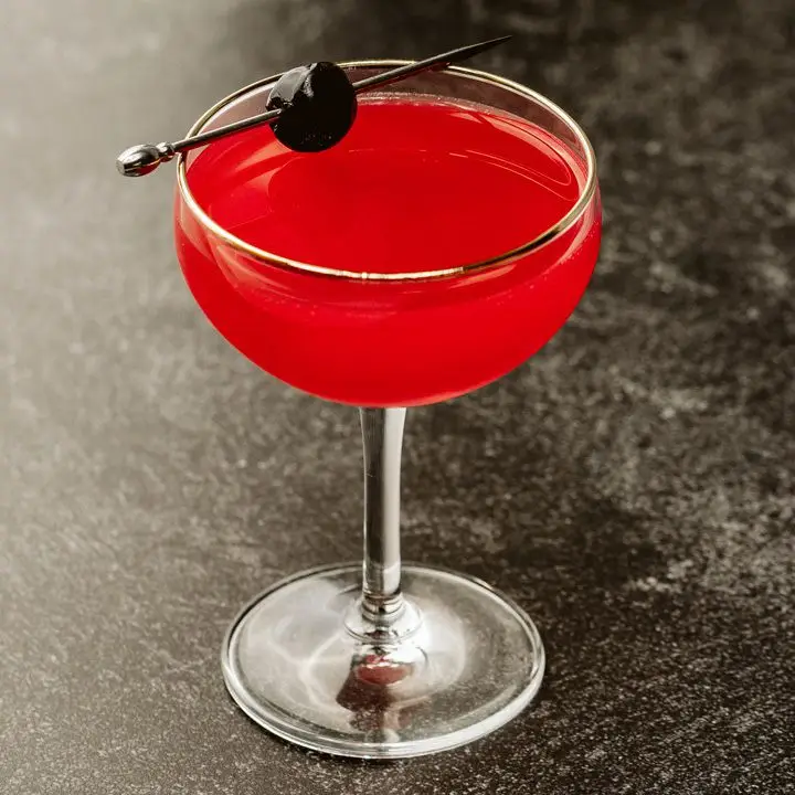 red-hued Modern cocktail in a coupe glass with a skewered cherry balanced on the rim