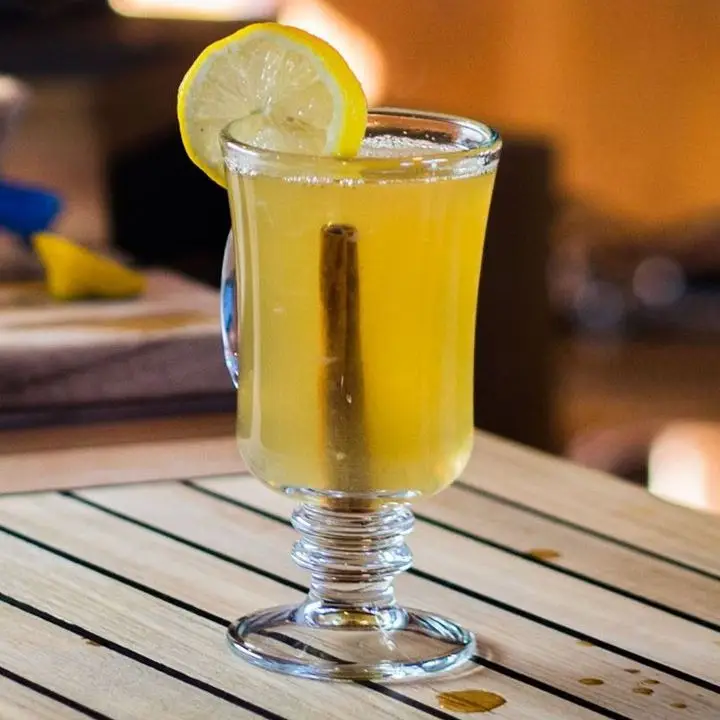 A Hot Toddy mugs rests on a slatted wooden table. It’s filled with a golden drink, a cinnamon stick floats at its base, and it is garnished with a lemon wheel.