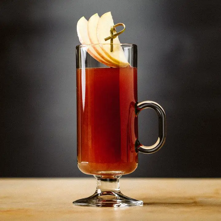 Autumn Wassail in a tall, thin toddy glass against a dark, slate-grey backdrop. A fan of three apple slices on a fancy skewer is the garnish. 